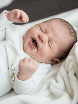 Crying newborn baby lying on a soft white blanket, wearing a white onesie.